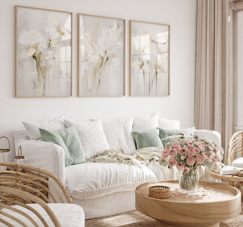 May include: Three framed abstract floral prints in muted tones of white and beige hang on a white wall above a white sofa with light green throw pillows. A vase of pink flowers sits on a wooden coffee table.