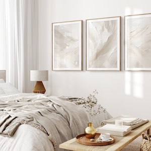 May include: Three framed abstract art prints in beige and white tones hang on a white wall above a bed with a white duvet and a beige throw blanket. A wooden side table with a lamp sits next to the bed. A wooden coffee table with a vase of dried flowers, a book, and a cup of coffee sits in front of the bed.