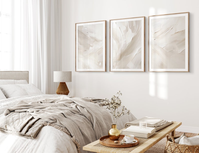 May include: Three framed abstract art prints in beige and white tones hang on a white wall above a bed with a white duvet and a beige throw blanket. A wooden side table with a lamp sits next to the bed. A wooden coffee table with a vase of dried flowers, a book, and a cup of coffee sits in front of the bed.