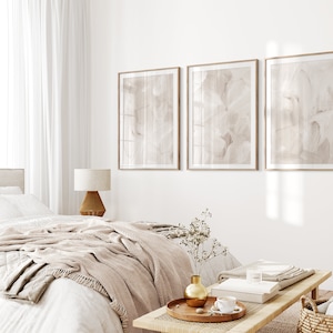 May include: A bedroom interior with a white bed, a beige throw blanket, and a wooden coffee table with a vase of dried flowers. Three framed abstract paintings in muted tones hang on the wall.