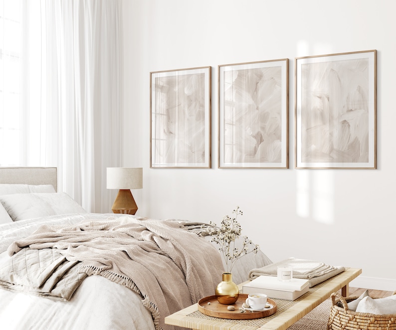 May include: A bedroom interior with a white bed, a beige throw blanket, and a wooden coffee table with a vase of dried flowers. Three framed abstract paintings in muted tones hang on the wall.
