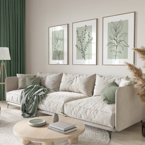 K&ouml;nnte beinhalten: Ein wei&szlig;es Sofa mit gr&uuml;nen Kissen und einer gr&uuml;nen Wolldecke steht in einem Wohnzimmer mit einem Holz-Couchtisch, einer Stehlampe und drei gerahmten Drucken von schwarzen Linienzeichnungen von Pflanzen an einer hellbeigen Wand.
