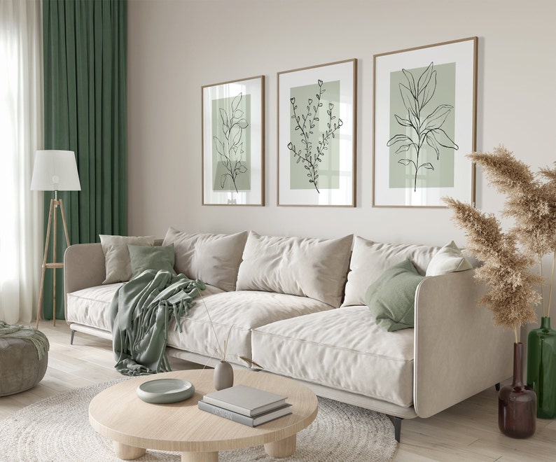 K&ouml;nnte beinhalten: Ein wei&szlig;es Sofa mit gr&uuml;nen Kissen und einer gr&uuml;nen Wolldecke steht in einem Wohnzimmer mit einem Holz-Couchtisch, einer Stehlampe und drei gerahmten Drucken von schwarzen Linienzeichnungen von Pflanzen an einer hellbeigen Wand.