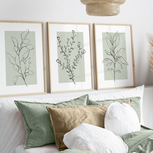 K&ouml;nnte beinhalten: Drei gerahmte Drucke mit minimalistischen Linienzeichnungen von Blatt&auml;sten auf einem Salbeigr&uuml;n-Hintergrund. Die Drucke sind in einer Reihe an einer wei&szlig;en Wand &uuml;ber einem Bett mit gr&uuml;nen und wei&szlig;en Kissen angeordnet.