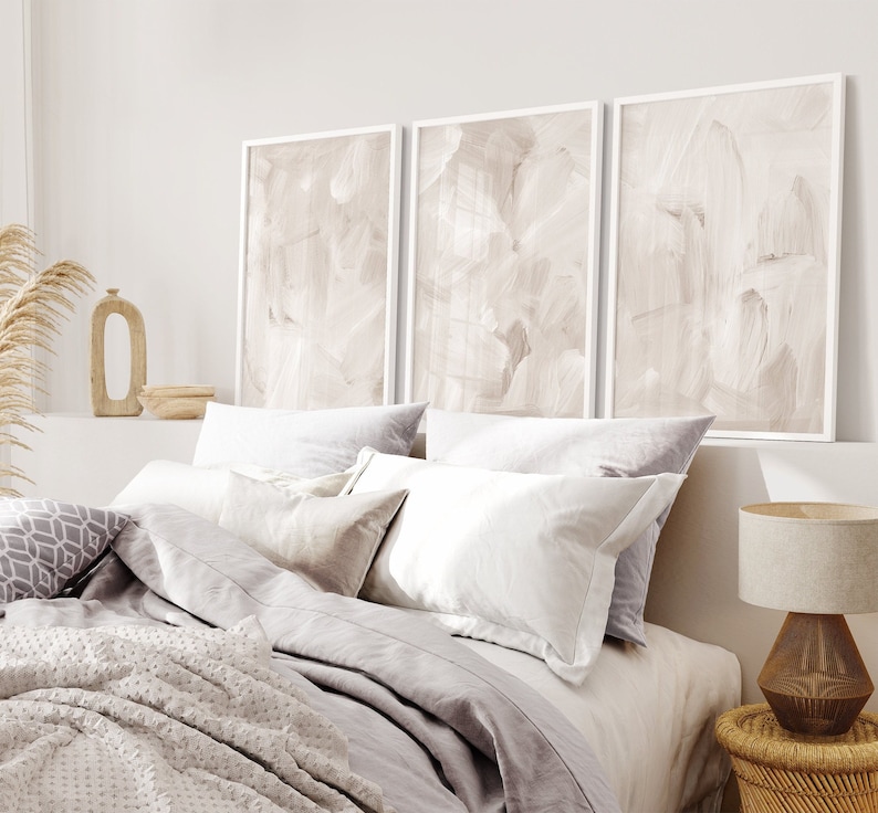 May include: A bedroom with a white bed with a grey duvet and white pillows. There are three abstract paintings above the bed with a light brown background. A small wooden table with a woven basket on top is in the corner of the room.