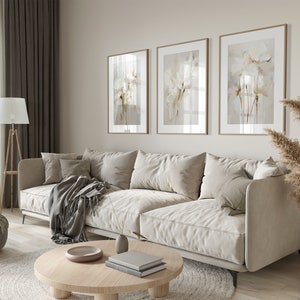May include: A light beige sectional sofa with three pillows and a gray throw blanket sits in a living room with a wooden coffee table, a round gray ottoman, and a floor lamp. Three framed floral prints hang on the wall behind the sofa. A vase with dried pampas grass sits on the floor next to the sofa.