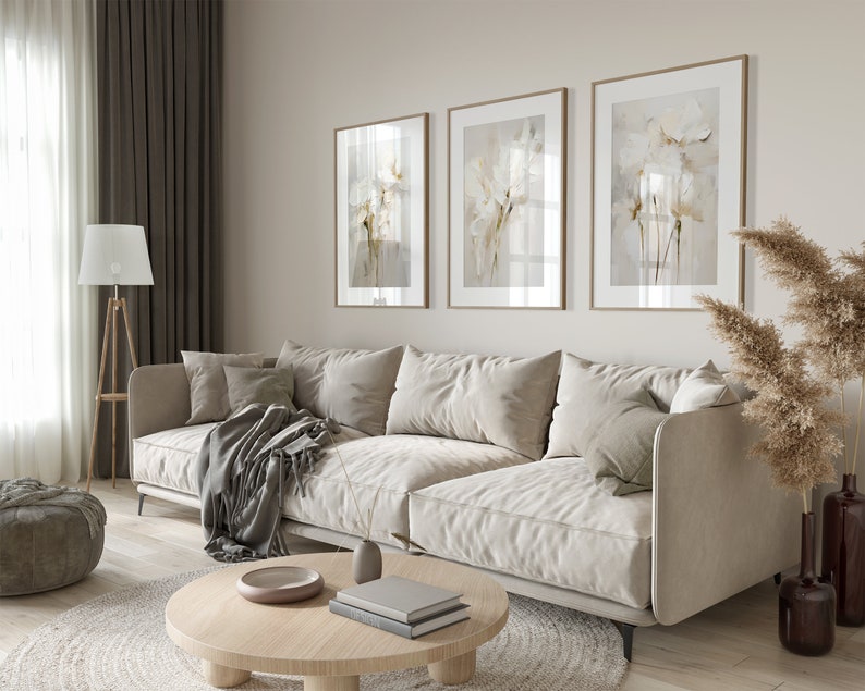 May include: A light beige sectional sofa with three pillows and a gray throw blanket sits in a living room with a wooden coffee table, a round gray ottoman, and a floor lamp. Three framed floral prints hang on the wall behind the sofa. A vase with dried pampas grass sits on the floor next to the sofa.
