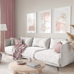May include: A white sectional sofa with pink throw pillows and a pink throw blanket in a living room with three framed prints of gold floral line art on a light beige wall.