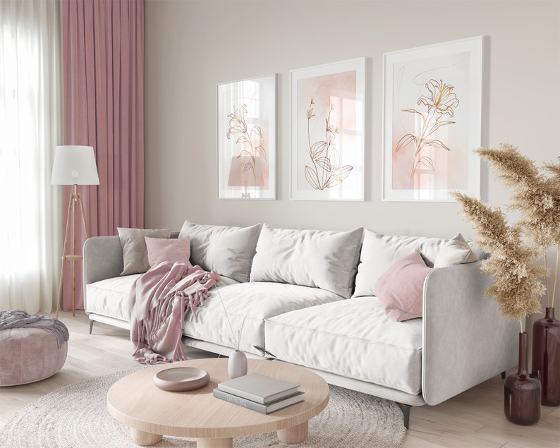 May include: A white sectional sofa with pink throw pillows and a pink throw blanket in a living room with three framed prints of gold floral line art on a light beige wall.