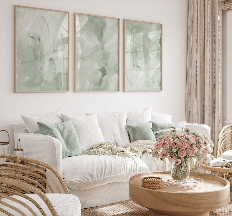 May include: A white sofa with green throw pillows and a blanket, a wooden coffee table with a vase of pink flowers, and three framed abstract paintings with green brushstrokes on a white wall.