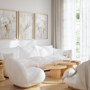 May include: A white sectional sofa with a white armchair and a set of two wooden nesting coffee tables in a living room with white walls and sheer curtains. The room is decorated with three framed floral paintings and a gold table lamp.