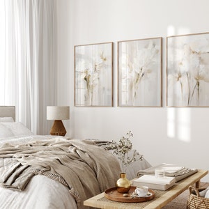 May include: Three framed abstract paintings with a white floral design on a light gray background. The paintings are hanging on a white wall in a bedroom setting with a bed, a lamp, and a side table.