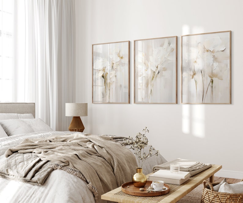 May include: Three framed abstract paintings with a white floral design on a light gray background. The paintings are hanging on a white wall in a bedroom setting with a bed, a lamp, and a side table.