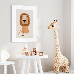 May include: A white framed print of a cartoon lion with a brown mane and a light brown body. The print is hanging on a white wall. There is a white table with a wooden stacking toy and wooden alphabet blocks on it. A white chair with bunny ears is next to the table. A large stuffed giraffe with brown spots is standing on the floor.