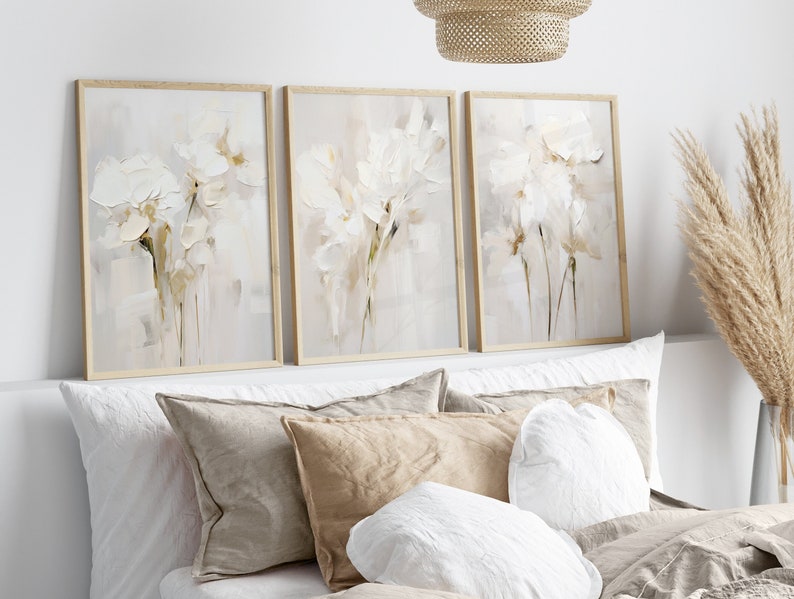 May include: Three framed abstract paintings with a white background and white flowers in shades of beige and brown.