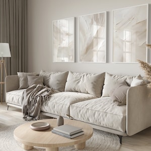 May include: A light beige sectional sofa with cushions and a throw blanket in a living room setting. The sofa is in front of a wall with three framed abstract art prints in beige tones. A round wooden coffee table with a bowl and books sits in front of the sofa. A dried pampas grass plant is in a vase on the right side of the image.