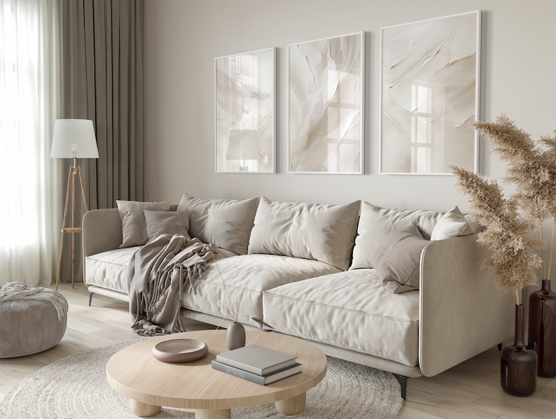 May include: A light beige sectional sofa with cushions and a throw blanket in a living room setting. The sofa is in front of a wall with three framed abstract art prints in beige tones. A round wooden coffee table with a bowl and books sits in front of the sofa. A dried pampas grass plant is in a vase on the right side of the image.