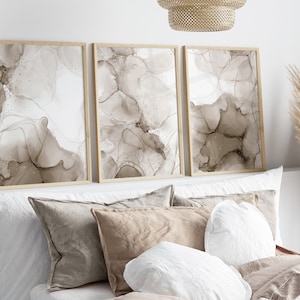 Puede incluir: Tres impresiones de arte abstracto enmarcadas con dise&ntilde;os de salpicaduras de tinta marr&oacute;n y blanca. Las impresiones est&aacute;n colgadas en una pared blanca sobre una cama con almohadas blancas y marrones.