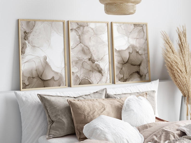 Puede incluir: Tres impresiones de arte abstracto enmarcadas con dise&ntilde;os de salpicaduras de tinta marr&oacute;n y blanca. Las impresiones est&aacute;n colgadas en una pared blanca sobre una cama con almohadas blancas y marrones.