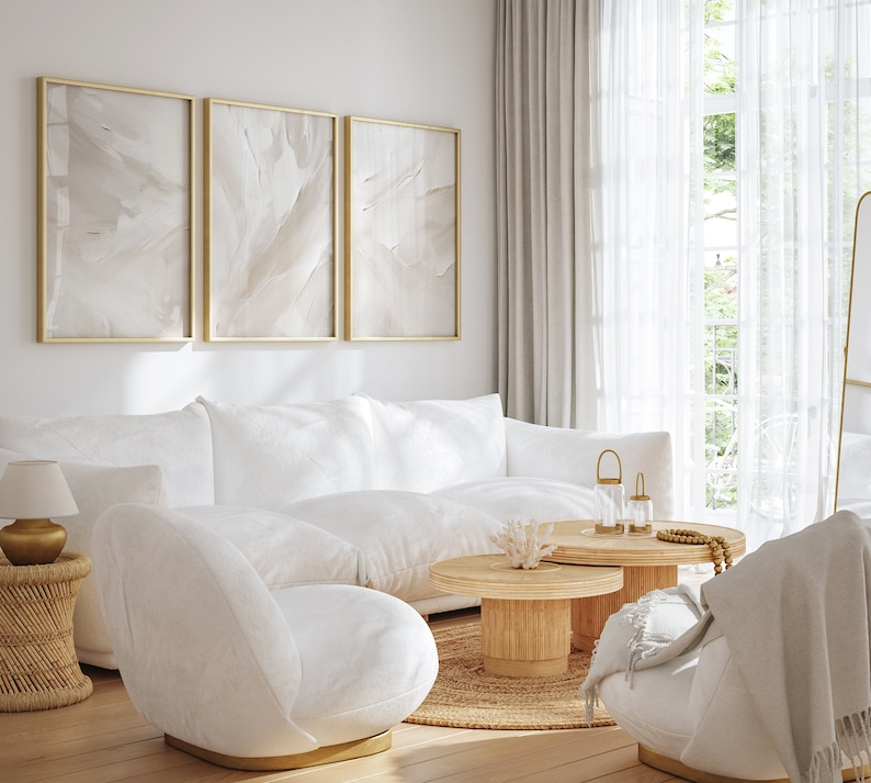 May include: A white living room with a white sofa, two white chairs, a wooden coffee table, and three framed abstract paintings on the wall. The room has a large window with white curtains and a mirror on the wall.