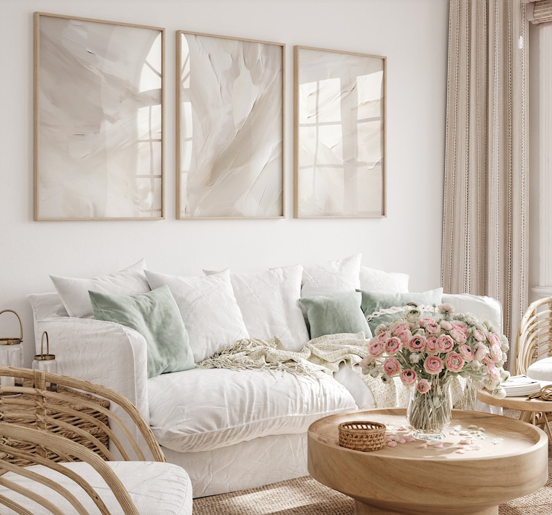May include: A white sofa with light green cushions and a white throw. A vase of pink flowers sits on a wooden coffee table in front of the sofa. Three framed abstract art prints hang on the wall behind the sofa.