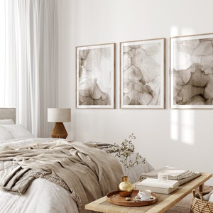 Puede incluir: Tres impresiones de arte abstracto en tonos marrones con fondo blanco, enmarcadas en madera natural, colgadas en una pared blanca sobre una cama con una manta beige y una mesita de noche de madera con un jarr&oacute;n y un libro.