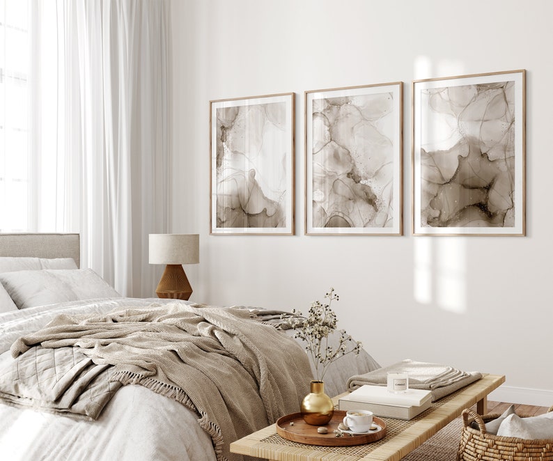 Puede incluir: Tres impresiones de arte abstracto en tonos marrones con fondo blanco, enmarcadas en madera natural, colgadas en una pared blanca sobre una cama con una manta beige y una mesita de noche de madera con un jarr&oacute;n y un libro.