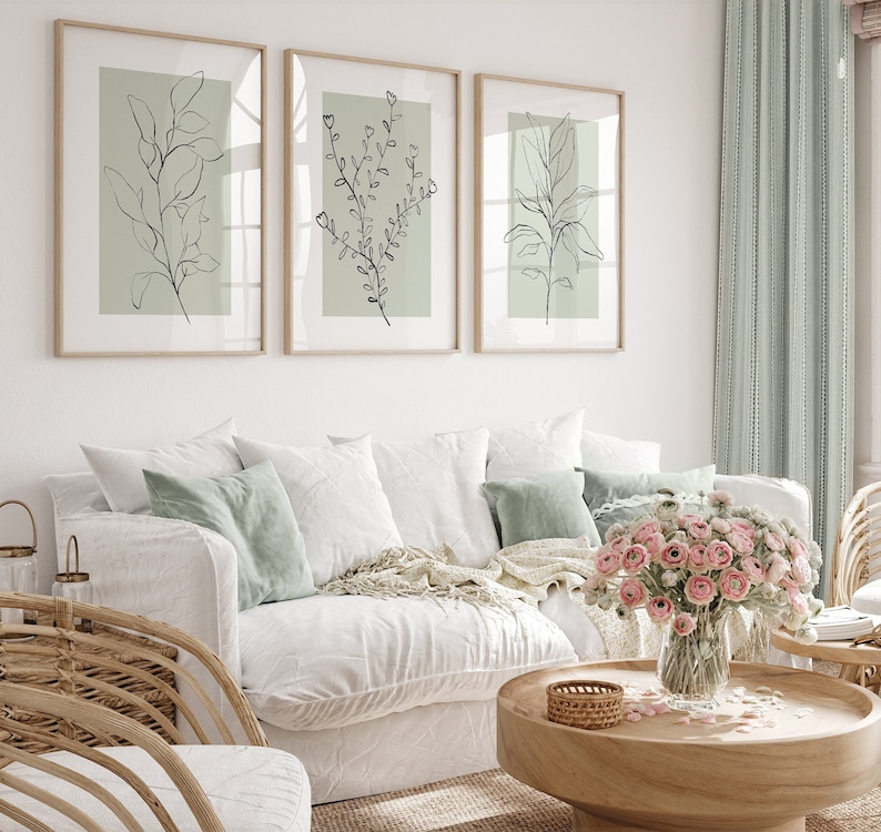 K&ouml;nnte beinhalten: Drei gerahmte Illustrationen von einfachen Strichzeichnungen von belaubten Zweigen auf hellgr&uuml;nem Hintergrund. Die Rahmen sind aus hellem Holz und h&auml;ngen an einer wei&szlig;en Wand &uuml;ber einem wei&szlig;en Sofa mit gr&uuml;nen Kissen. Ein Strau&szlig; rosa Blumen steht auf einem Holz-Couchtisch vor dem Sofa.