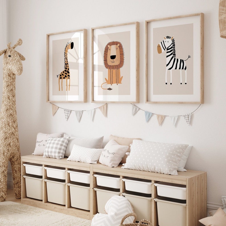 May include: A white wall with three framed animal prints. The prints feature a giraffe, a lion, and a zebra. The prints are hung above a wooden storage bench with white bins. A stuffed giraffe toy is on the left side of the image.