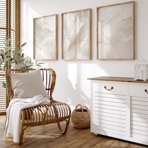 May include: A white wicker chair with a beige throw blanket and cushion sits in a room with three framed abstract prints on the wall and a white cabinet with a wood top.