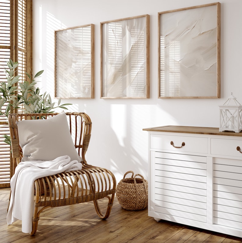 May include: A white wicker chair with a beige throw blanket and cushion sits in a room with three framed abstract prints on the wall and a white cabinet with a wood top.