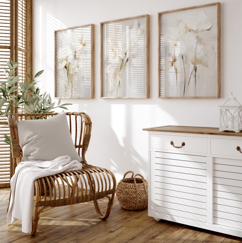 May include: A white wicker chair with a white throw blanket and a white pillow sits in a room with three framed floral prints on the wall and a white cabinet with a wooden top.