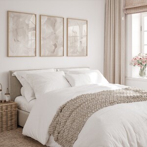 May include: A white bed with a beige knitted throw blanket and white pillows. The bed is against a white wall with three framed abstract paintings in beige tones. The room has a window with beige curtains and a vase of pink flowers.