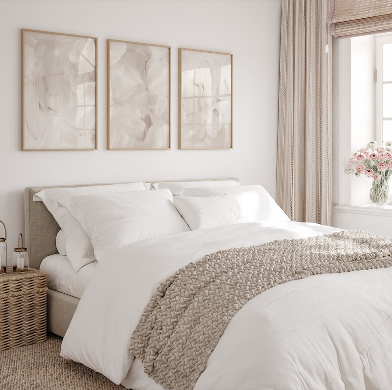 May include: A white bed with a beige knitted throw blanket and white pillows. The bed is against a white wall with three framed abstract paintings in beige tones. The room has a window with beige curtains and a vase of pink flowers.