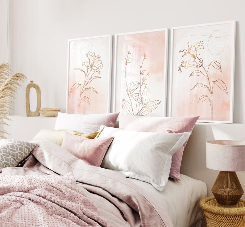May include: Three framed prints with a pink watercolor background and gold line drawings of flowers. The prints are hanging on a white wall above a bed with pink bedding.