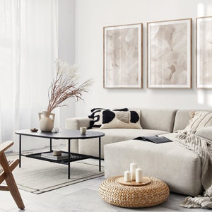May include: A modern living room featuring a large sectional sofa, a coffee table, and a woven ottoman. The sofa is upholstered in a light grey fabric and has several throw cushions. The coffee table is made of black metal with a glass top. The ottoman is round and has a woven top. Three framed abstract paintings adorn the wall behind the sofa.