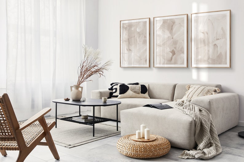 May include: A modern living room featuring a large sectional sofa, a coffee table, and a woven ottoman. The sofa is upholstered in a light grey fabric and has several throw cushions. The coffee table is made of black metal with a glass top. The ottoman is round and has a woven top. Three framed abstract paintings adorn the wall behind the sofa.