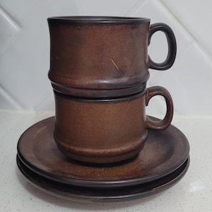 May include: Two stacked brown ceramic mugs with matching saucers. The mugs feature a cylindrical shape with a rounded base and a small handle on the side. The ceramic has a dark, mottled brown finish with a smooth texture and a subtle sheen.