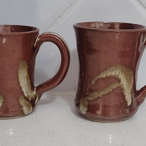 May include: Two handmade ceramic mugs with a glossy reddish-brown glaze and irregular, textured beige brushstroke patterns. Each mug features a wide, flared rim, a sturdy curved handle, and a cylindrical body with a slightly tapered base design.