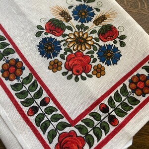 May include: White linen napkin with a floral design. The center features a bouquet of red roses, blue flowers, and wheat. A red border with green leaves and berries frames the edges. The napkin is folded.