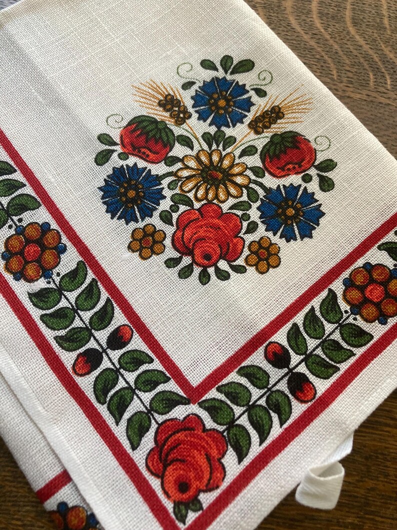 May include: White linen napkin with a floral design. The center features a bouquet of red roses, blue flowers, and wheat. A red border with green leaves and berries frames the edges. The napkin is folded.