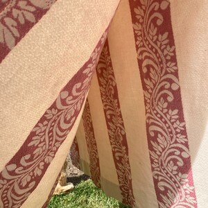 May include: A fabric structure featuring alternating vertical panels of cream-colored material and wide, deep red bands decorated with a repeating intricate floral and leaf scroll pattern. The textile hangs in folds, creating a tent-like opening.