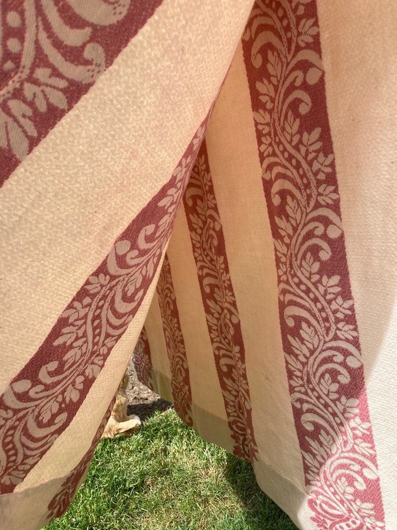 May include: A fabric structure featuring alternating vertical panels of cream-colored material and wide, deep red bands decorated with a repeating intricate floral and leaf scroll pattern. The textile hangs in folds, creating a tent-like opening.