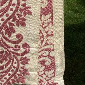 May include: A textile fabric featuring a cream-colored base with a woven herringbone texture. The material is decorated with a deep red damask-style pattern consisting of intricate floral and leaf motifs, with a distinct vertical border design.