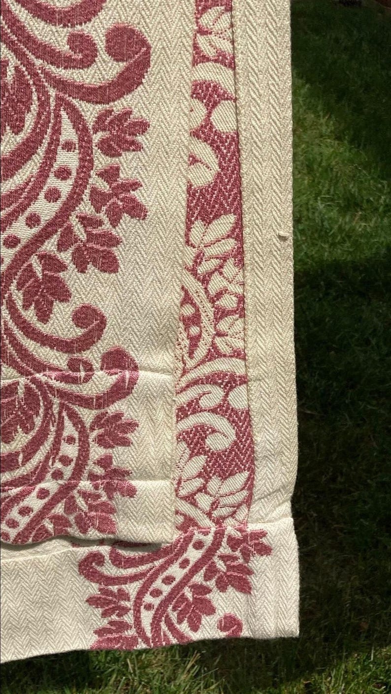 May include: A textile fabric featuring a cream-colored base with a woven herringbone texture. The material is decorated with a deep red damask-style pattern consisting of intricate floral and leaf motifs, with a distinct vertical border design.