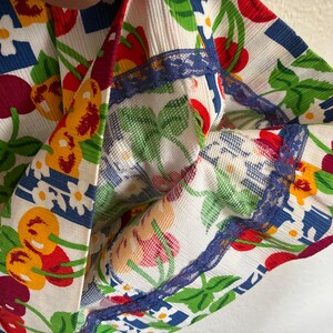 K&ouml;nnte beinhalten: Nahaufnahme eines Stoffes mit einem lebendigen Kirschmuster in Rot, Gelb und Lila, vor einem wei&szlig;en Hintergrund. Das Design umfasst gr&uuml;ne Bl&auml;tter, wei&szlig;e Blumen und blaue geometrische Formen. Die Kanten sind mit blauer Spitze verziert.