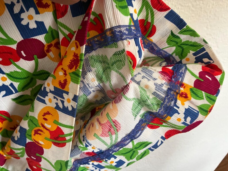 K&ouml;nnte beinhalten: Nahaufnahme eines Stoffes mit einem lebendigen Kirschmuster in Rot, Gelb und Lila, vor einem wei&szlig;en Hintergrund. Das Design umfasst gr&uuml;ne Bl&auml;tter, wei&szlig;e Blumen und blaue geometrische Formen. Die Kanten sind mit blauer Spitze verziert.