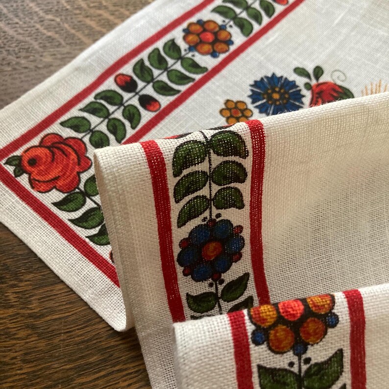 May include: Close-up of white linen fabric with a red border featuring floral designs. The designs include red roses, blue and yellow flowers, and green leaves. The fabric is folded, showing the pattern on the edges.