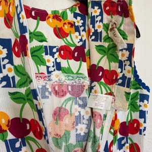 K&ouml;nnte beinhalten: &Auml;rmelloses Kleidungsst&uuml;ck im Vintage-Stil mit wei&szlig;em Hintergrund, verziert mit einem lebendigen Kirsch- und Blumenmuster in Rot, Gelb und Gr&uuml;n. Das Kleidungsst&uuml;ck hat einen Rei&szlig;verschluss und ist mit blauer und wei&szlig;er Spitze verziert.