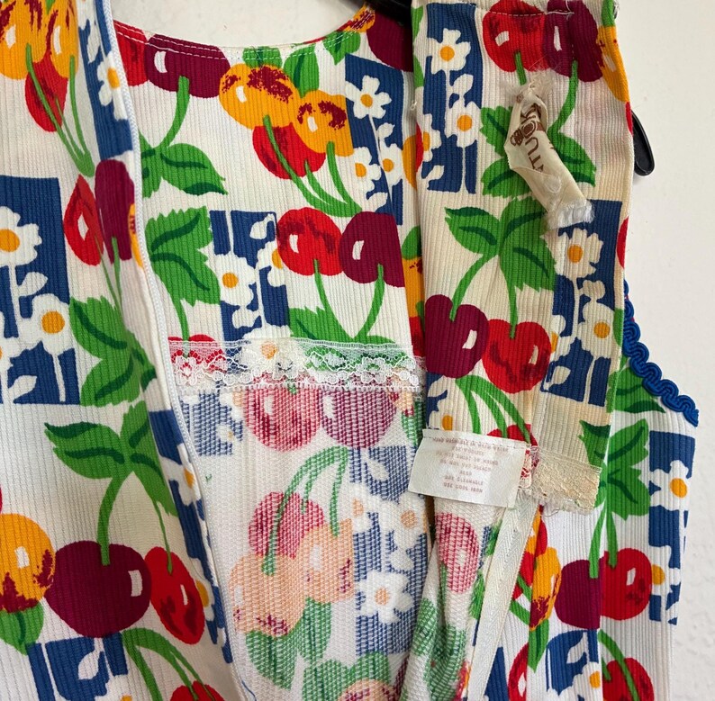 K&ouml;nnte beinhalten: &Auml;rmelloses Kleidungsst&uuml;ck im Vintage-Stil mit wei&szlig;em Hintergrund, verziert mit einem lebendigen Kirsch- und Blumenmuster in Rot, Gelb und Gr&uuml;n. Das Kleidungsst&uuml;ck hat einen Rei&szlig;verschluss und ist mit blauer und wei&szlig;er Spitze verziert.